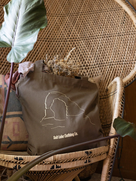 Desert Tote bag in brown canvas with Salt Lake Clothing label, displayed against a woven chair, holding dried flowers and surrounded by plants and earthy home decor.